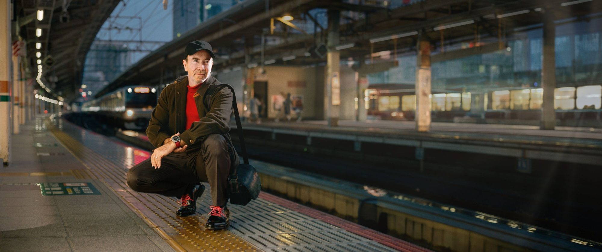 Laurence Balter crouching on a Tokyo train platform at dusk with a leather Oberwerth bag over his shoulder