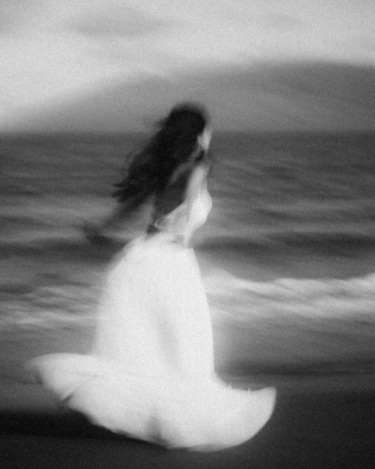 Black and white motion blur of a figure in a white dress at the water&rsquo;s edge