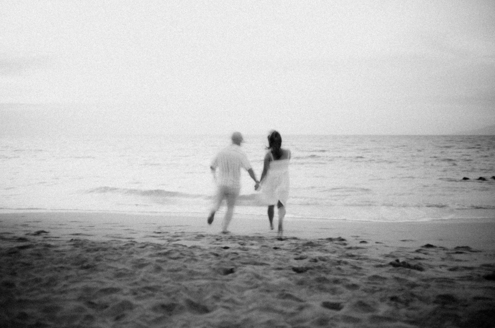 Black and white long exposure of a couple running hand in hand toward the sea