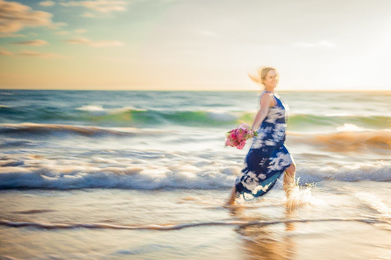 Color long exposure of a woman in a blue dress carrying a bouquet through shallow surf at sunset