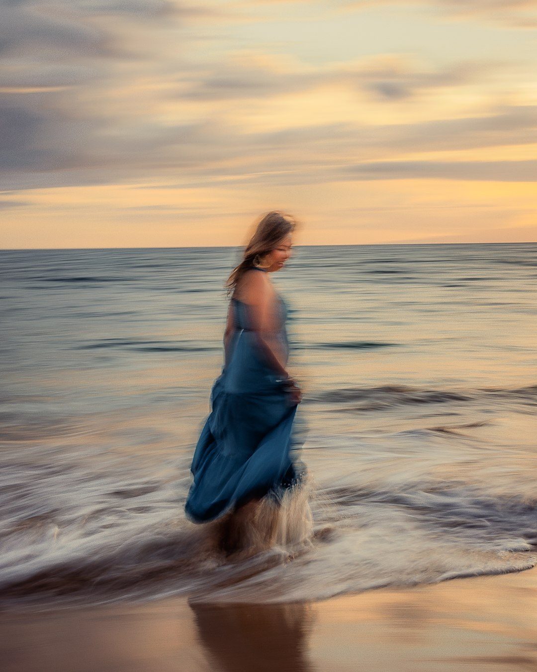 Color long exposure of a woman in a blue dress walking in the surf at sunset