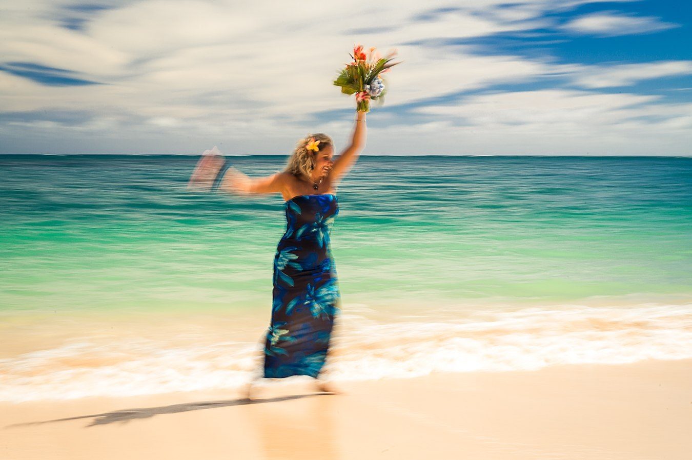 Color long exposure of a woman in a dark floral dress raising a bouquet before turquoise water