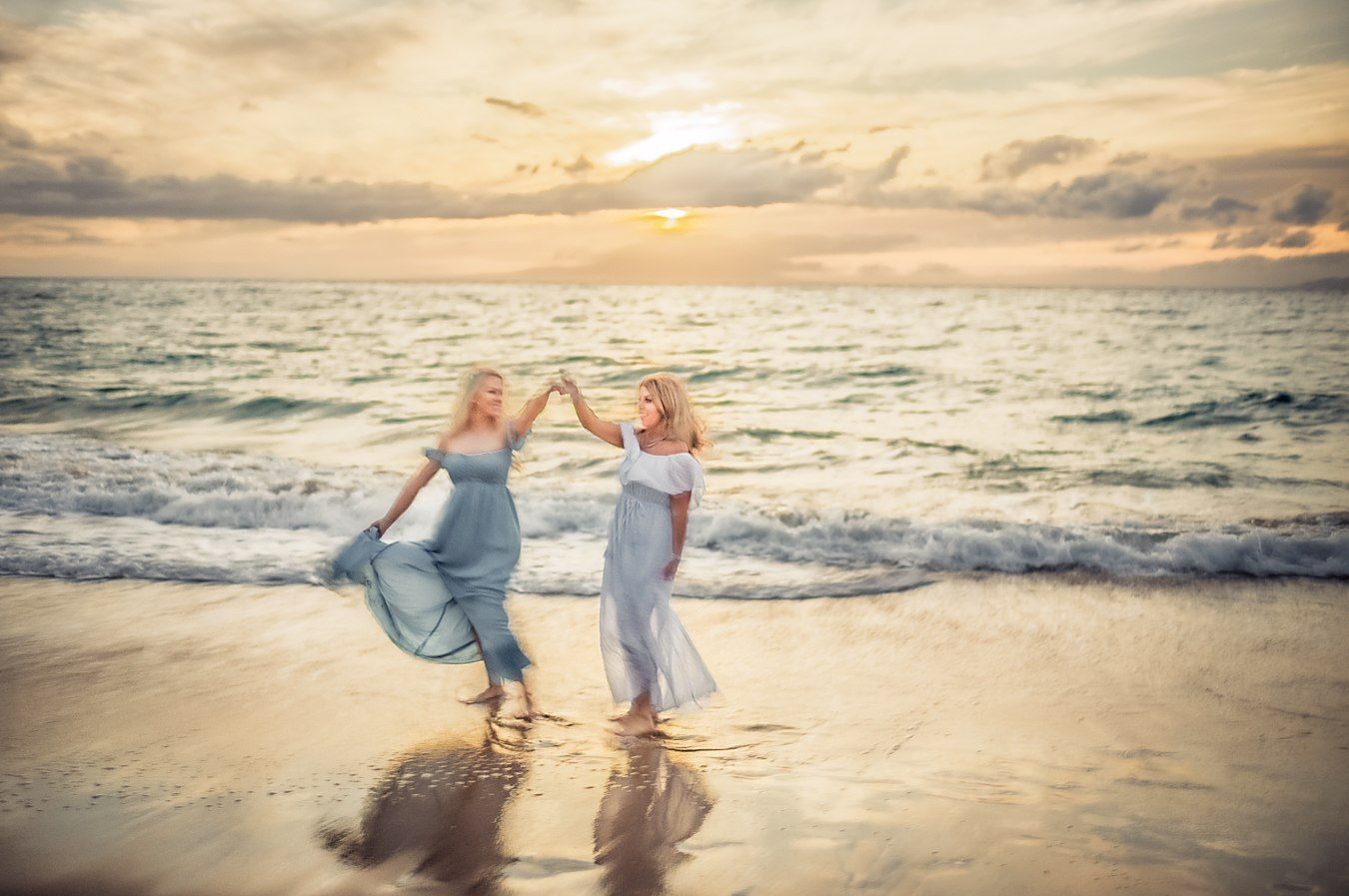 Color photograph of two women in pale dresses dancing at the water&rsquo;s edge at sunset