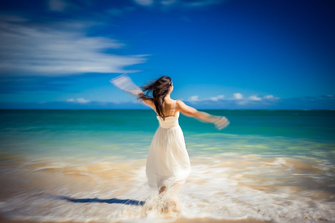 Color long exposure of a woman in a white dress with arms outstretched before a deep blue sky and teal sea