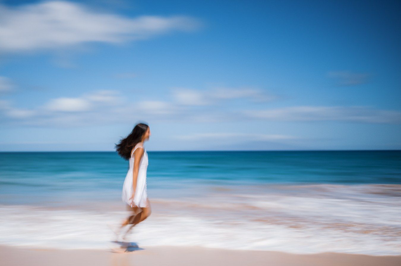 Color long exposure of a woman in a white dress walking past a streaked blue sea and sky