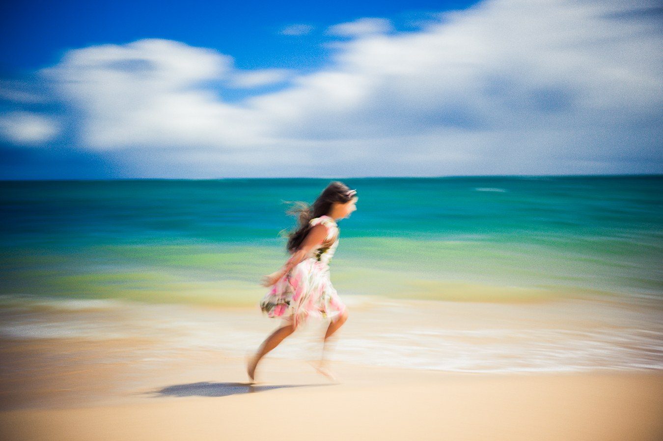 Color long exposure of a girl in a floral dress running along the shore beside a teal sea