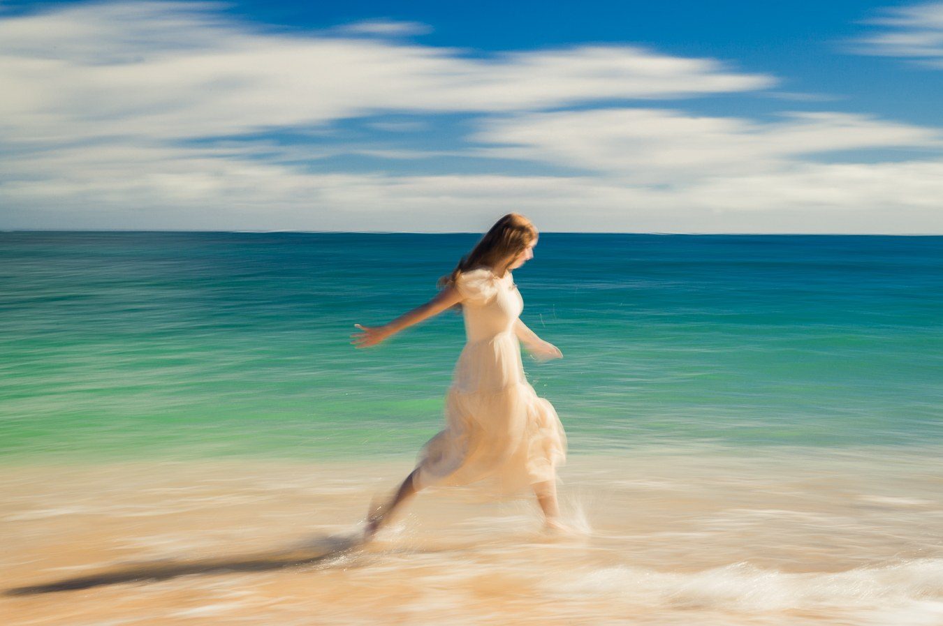 Color long exposure of a woman in a cream dress running through the surf beside a teal sea