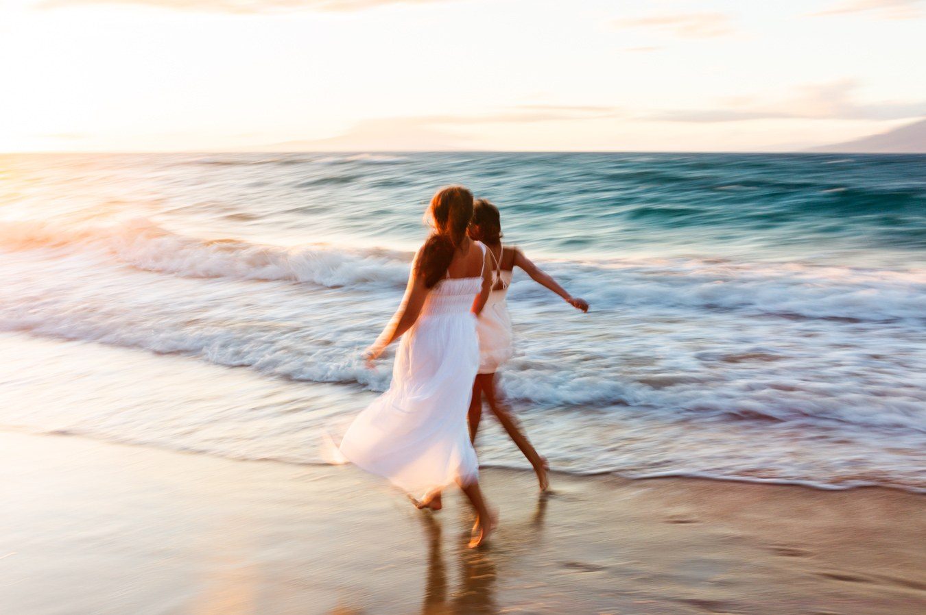 Color long exposure of two figures in white dresses running along the shore at sunset