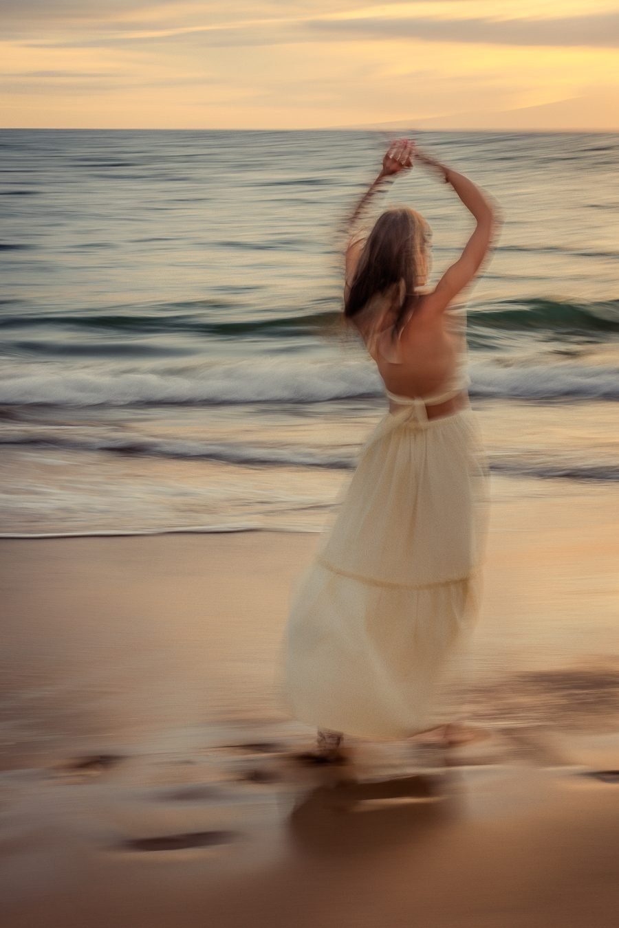 Color long exposure of a woman in a cream dress with arms raised, dancing on the beach at dusk