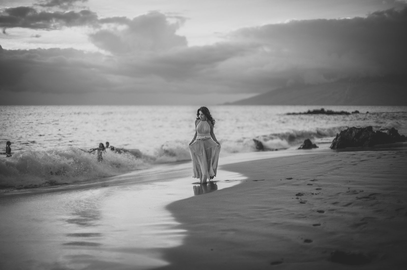 Black and white portrait of a woman in a flowing dress walking along the water&rsquo;s edge as waves break behind her